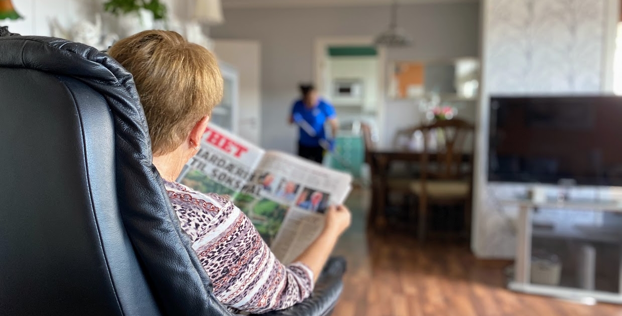 Bruker leser avis i front, samtidig som hjemmehjelp vasker i bakgrunnen. 